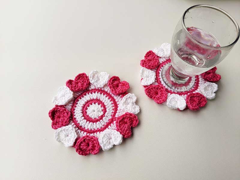 two crochet coasters with decor hearts laying next to each other with a glass of water standing on the top of one of them