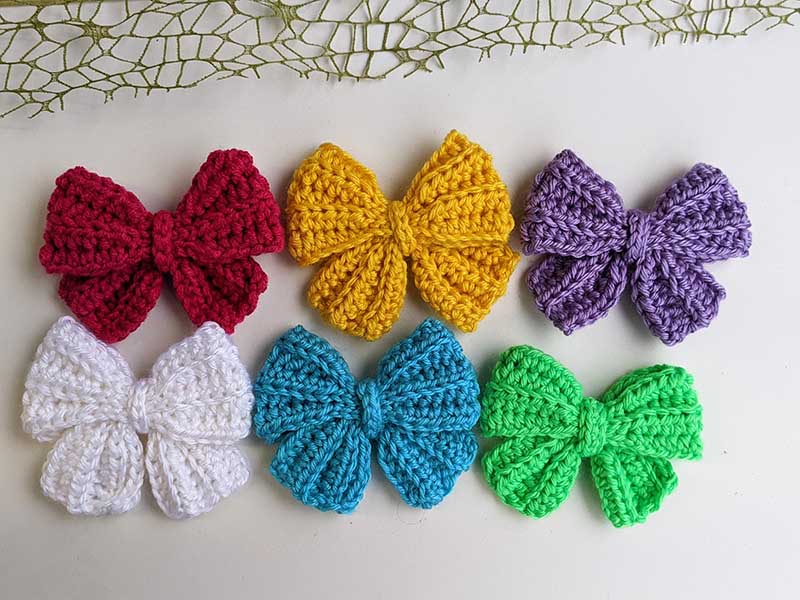 six crochet hair bows arranged in two rows. red, yellow, and purple bows are in the first row whereas white, blue, and green bows are in the second row.
