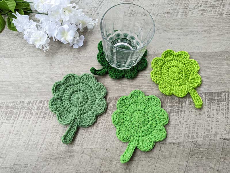 four crochet shamrock or clover leaf coasters made in different shades of green color and glass of water standing on one of them