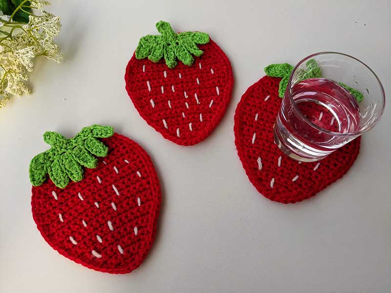 three crochet strawberry coasters next to each other with a glass of water on one of them