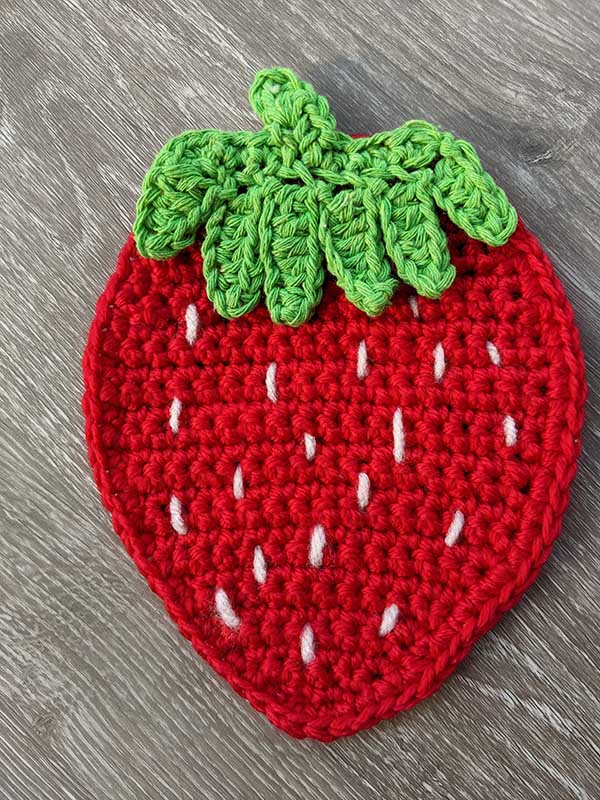 crochet strawberry coaster with white embroidered seeds and green leaves