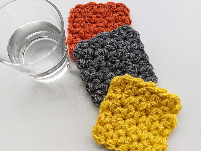 three crochet jasmine stitch coasters made in red/orange, dark gray, and bright yellow yarn
